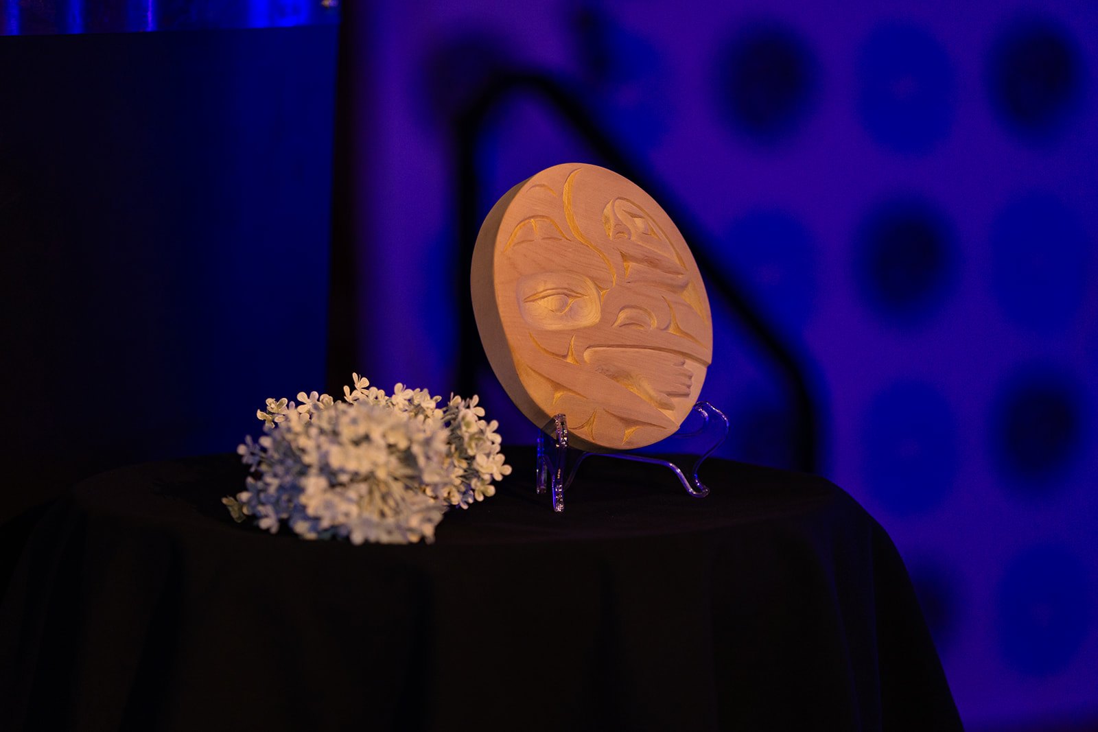 A round wooden carving of the artwork on a clear stand beside white flowers, lit in blue.