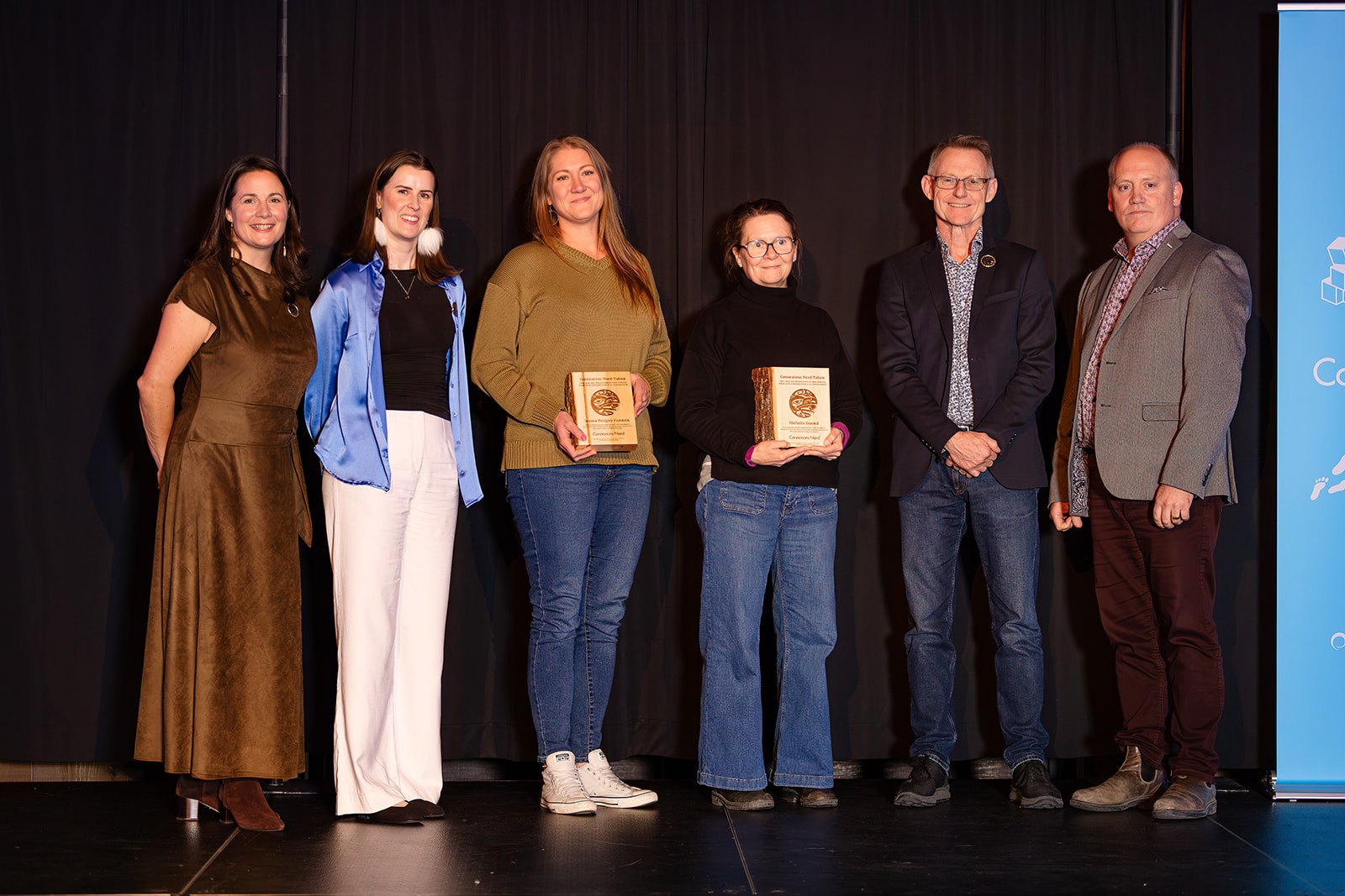 Six people on stage; two hold wooden award plaques.