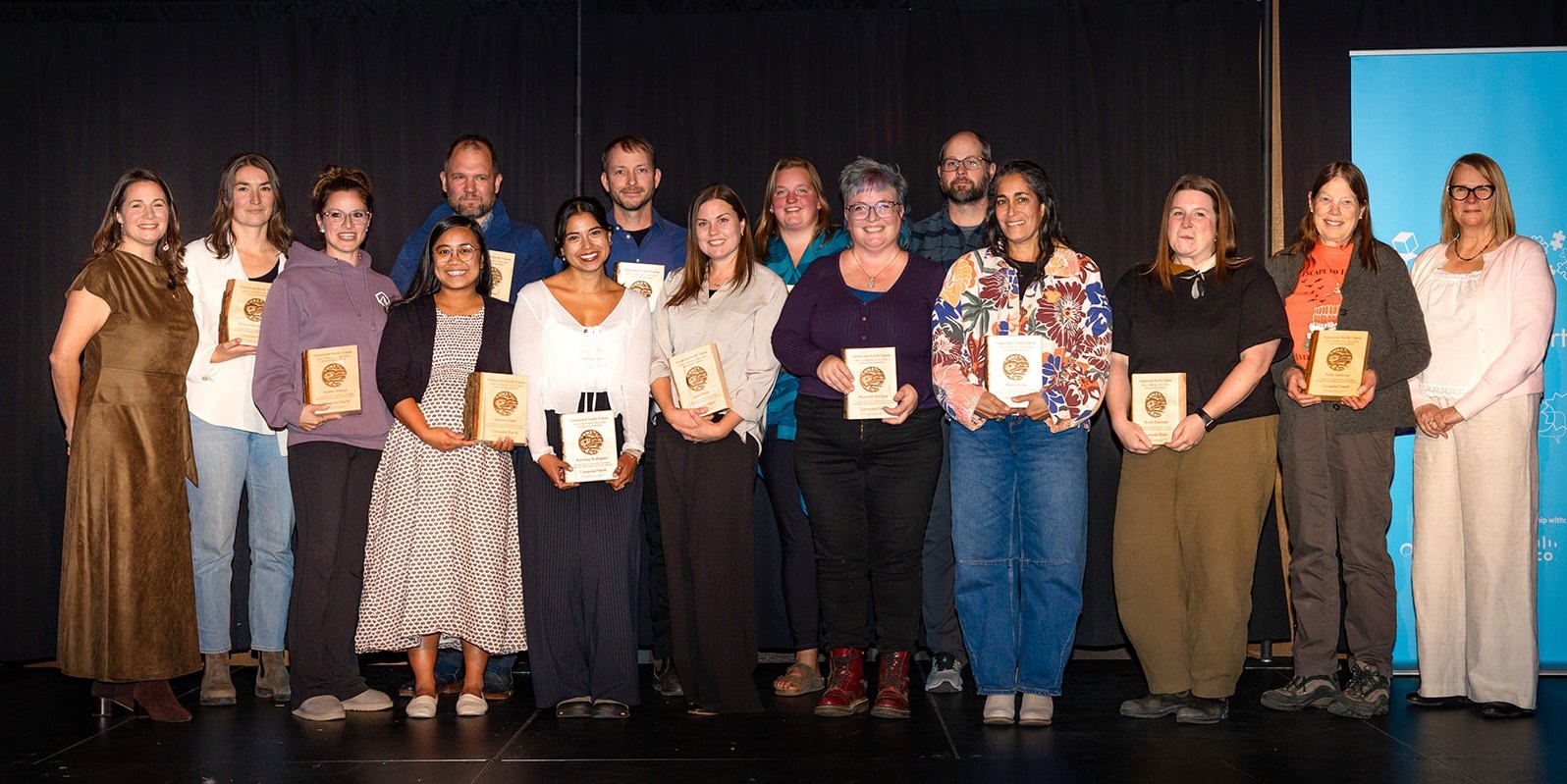 Educators stand in two rows on stage holding wooden award plaques.