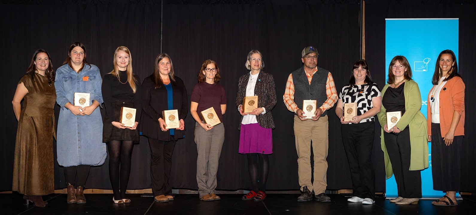 First Nation School Board school awardees stand in a row on stage, most holding wooden award plaques.